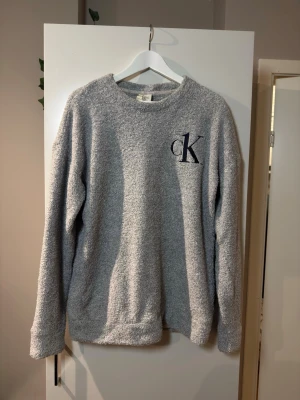 Grå teddytröja från Calvin Klein - Mysig grå teddytröja från Calvin Klein med broderad CK-logga på bröstet. Tröjan har rund halsringning, långa ärmar och är gjord i ett fluffigt, mjukt material som känns riktigt skönt mot huden. Perfekt för chill dagar hemma.