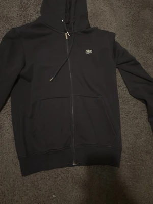 Svart zip-hoodie från Lacoste - Snygg svart hoodie från Lacoste med dragkedja framtill och klassisk krokodillogga på bröstet. Hoodien har huva med dragsko och två stora fickor framtill. Perfekt för en avslappnad och stilren look. Tar helst imot kontanter och kan mötas upp Stockholm