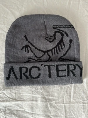 Grå mössa från Arc'teryx - Snygg grå mössa från Arc'teryx med svart logotyp och grafiskt mönster framtill. Mössan är ribbstickad med uppvikt kant och har en modern, clean look. Perfekt för kalla dagar och passar till de flesta outfits.