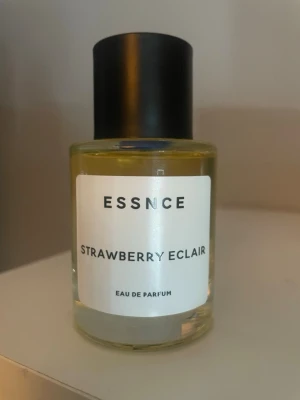 ESSNCE Strawberry Eclair parfym - ESSNCE Strawberry Eclair Eau de Parfum i en rund glasflaska med svart lock. Parfymen har en ljusgul vätska och en stilren vit etikett med svart text. Doften har troligen söta och fruktiga toner inspirerade av jordgubb och bakverk.