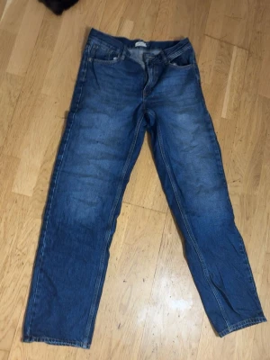 Blåa lågmidjade jeans - Säljer ett par klassiska blå jeans med rak passfor. Materialet är denim  och färgen är mellanblå med lätt slitning på låren. Perfekta till en avslappnad stil.