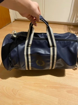 Mörkblå barrel bag från Fred Perry - Snygg mörkblå barrel bag från Fred Perry i konstläder med vita och svarta ränder på handtagen. Väskan har ett stort huvudfack, sidofack och justerbar axelrem. Guldigt logotryck på sidan och klassisk sportig look. Använder  några gånger.  Pris kan diskuteras 