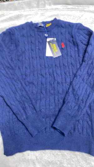 Blå kabelstickad tröja Polo Ralph Lauren - Snygg blå kabelstickad tröja från Polo Ralph Lauren med rund halsringning och röd broderad logga på bröstet. Tröjan har långa ärmar och ribbade muddar vid ärmslut och nederkant. Perfekt för dig som gillar klassisk och stilren design.