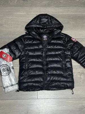 Svart dunjacka från Canada Goose - Säljer en svart dunjacka från Canada Goose med huva och två stora dragkedjeförsedda fickor framtill. Jackan har det klassiska Canada Goose-märket på ärmen och är tillverkad i glansigt material. Perfekt för kalla vinterdagar och riktigt snygg streetstil.