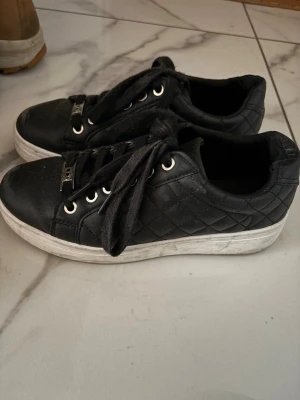 Svarta quiltade sneakers med vita sulor - Snygga svarta sneakers med quiltat mönster på sidorna och vita, tjocka sulor. Skorna har svarta skosnören, silverfärgade öljetter och dekorativa metalldetaljer framtill. Perfekta för en trendig och avslappnad look.