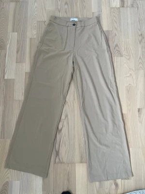 Beiga vida kostymbyxor - Säljer ett par beiga kostymbyxor från Bershka med hög midja och vida ben. Byxorna har en klassisk knappstängning fram, pressveck och en bakficka med knapp. Materialet känns mjukt och har en lätt stretch för extra komfort. Innebenslängden är ungefär 78cm