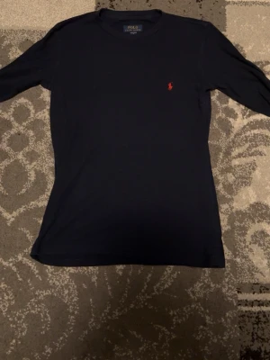 Mörkblå långärmad tröja Polo Ralph Lauren - Mörkblå långärmad tröja från Polo Ralph Lauren med klassisk röd broderad logga på bröstet. Tröjan har rund halsringning och är tillverkad i mjukt material som sitter skönt på kroppen. Perfekt för en clean och stilren look. Storlek S sitter som en lite större S