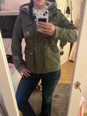 Grön parkajacka med broderier - Snygg grön parkajacka med huva och broderade detaljer på ärmar, bröst och axel. Jackan har flera stora fickor framtill, dragkedja och tryckknappar. Insidan är fodrad med mjuk teddy och jackan har en cool patch på ena ärmen.