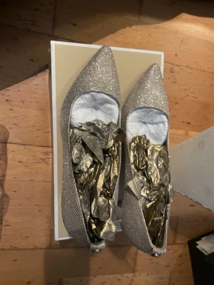 Michael Kors Glittriga silvriga pumps  - Säljer ett par glittriga silvriga pumps med spetsig tå och låg klack. Skorna har en elegant form och är täckta av skimrande material som fångar ljuset. Perfekta för att lyfta vilken outfit som helst. Inget synligt märke på skorna