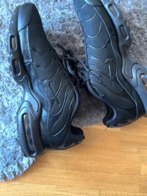 Nike Air Max Plus helsvarta sneakers - Säljer ett par helsvarta Nike Air Max Plus sneakers med mesh- och syntetmaterial, ikoniska vågmönster och synlig Air-dämpning i sulan. Skorna har lågt skaft och klassisk snörning. Perfekta för dig som gillar streetwear och sportig stil.