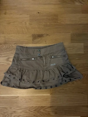 Beige volangkjol med dragkedjor från dcp - Säljer en beige kortkjol från dcp med volangkant och metallöljetter längs nederdelen. Kjolen har två dragkedjefickor framtill och dubbla knappar i midjan. Perfekt för en trendig och avslappnad stil. Midjemåttet är 33cm💕💕