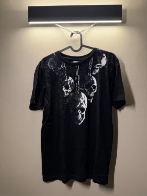 Svart t-shirt med dödskalletryck - Cool svart t-shirt från Indiska med stort tryck av kedjor och dödskallar på bröstet. Klassisk rund halsringning och korta ärmar. Perfekt för dig som gillar edgy och alternativ stil.
