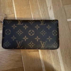 Plånbok  - Helt ny plånbok med mönster. Även Mussepigg på en sida. Fack för kort, sedlar och fack för mynt med dragkedja. Följer med en sling, så man kan bära den på handleden också, glömde fota den, skriv om ni vill se den. Är en svart sling. Följer med dustbag och box till också. Kan skicka bild på dom också. Lapp sitter kvar också. Är ca 19-20cm bred och va 11-12cm hög