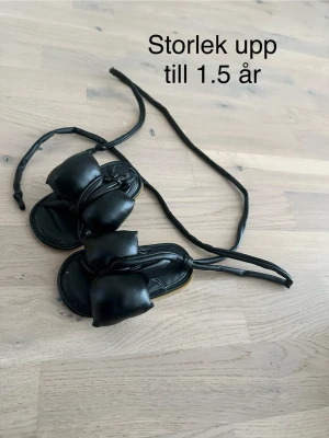 Svarta babysandaler med knytband - Säljer ett par svarta sandaler för baby, med mjuka knytband runt ankeln och vadderade detaljer över foten. Sandalerna är tillverkade i konstläder och passar upp till 1,5 år. Perfekta för små fötter som vill ha något bekvämt och stilrent.