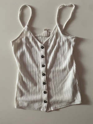 Vit ribbad linnetopp från H&M - Säljer en vit ribbad linnetopp från H&M med smala axelband och svarta knappar framtill. Toppen har v-ringning och är tillverkad i mjuk bomullsmix. Tröjan är tunn och därför kan man behöva något under. 