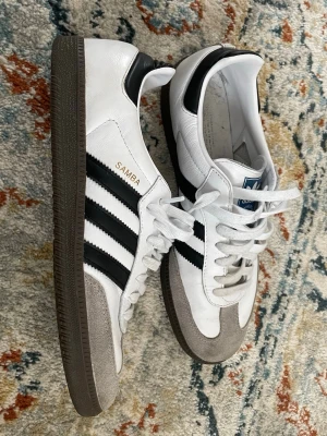Adidas samba  - Klassiska Adidas Samba sneakers i vitt läder med svarta ränder och detaljer. Skorna har grå mocka framtill och mörkbrun gummisula. Ikonisk design med låg profil och snörning. Använd 3 gånger