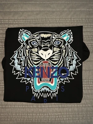 Svart Kenzo t-shirt med tigertryck - Snygg svart t-shirt från Kenzo med stort broderat tigerhuvud i vitt, blått och turkost på bröstet. Klassisk passform och rund hals. Kenzo Paris-loggan är broderad i blått över motivet. Perfekt statement-plagg för dig som gillar streetwear.