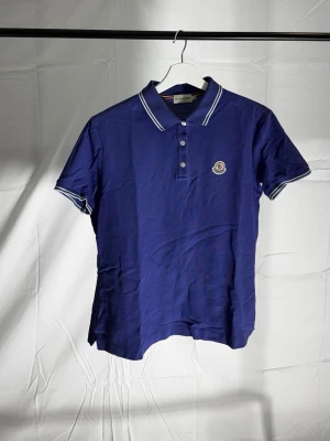 Blå pikétröja från Moncler - Sick 8.5/10.                 Bild:6 ser vi en liten skada som är minimal som inte syns om man inte vet att den finns där. Annars är piken bra sick och väldigt fin. Storleken är EU S men sitter lite mindre än S men inte nära XS.                                                         Snygg blå pikétröja från Moncler med vit kontrastkant på krage och ärmslut. Tröjan har korta ärmar, knappslå med tre knappar och Moncler-logga på bröstet. Tillverkad i mjukt bomullsmaterial som känns skönt mot huden.