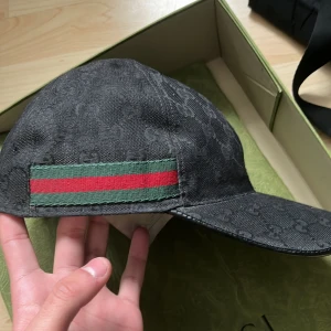 Svart keps från Gucci (äkta) - Säljer en svart keps från Gucci med klassiskt GG-monogram över hela kepsen som är äkta. På sidan finns en grön och röd rand som signaturdetalj. Kepsen har böjd skärm och är tillverkad i ett slitstarkt tygmaterial. Perfekt för dig som gillar exklusiva accessoarer. Gucci lappen har tappat färg så det är svårt o se vad som står.
