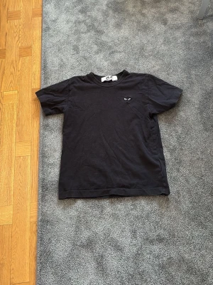 Svart Comme des Garçons t-shirt - Säljer en svart t-shirt från Comme des Garçons Play med det ikoniska hjärta med ögon broderat på bröstet. Klassisk passform och rund halsringning. Perfekt basic-plagg med en stilren twist.