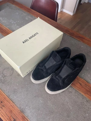 Svarta Axel Arigato slip-on sneakers - Stilrena svarta slip-on sneakers från Axel Arigato med vit sula. Skorna har en minimalistisk design utan snörning och är tillverkade i mocka och textil. Perfekta för en clean och modern look.
