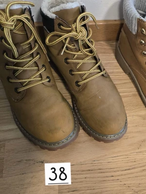 Bruna kängor med foder och snörning - Snygga bruna kängor med gulaktiga snören och grov sula. Insidan är fodrad med vitt, mjukt material för extra värme. Kängorna har metallhål för snörningen och en klassisk outdoor-look. Perfekta för kalla dagar och streetstyle.