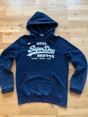 Superdry Vintage hoodie - Säljer nu en mörkblå hoodie från Superdry med stor vit logga och japansk text på bröstet. Klassisk känguruficka framtill, ribbade muddar och nederkant samt justerbar huva med dragsko. Tillverkad i mjukt bomullsmaterial. Tröjan är i bra skick och har inga defekter.   Mått: Längd från axel 70 cm. Bredd armhåla till armhåla 55 cm.  Om ni har någon fundering tveka inte att höra av er! 😊