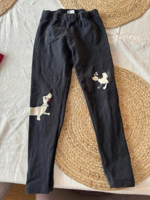Svarta leggings med hundmotiv - Säljer ett par svarta leggings med gulliga hundmotiv på benen. De har resår i midjan och är gjorda i mjukt stretchigt material som sitter skönt. Storlek 104