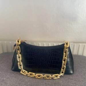 Svart axelväska med guld kedja - Stilren svart axelväska i croco-mönstrat konstläder med en chunky guldfärgad kedja som axelrem. Väskan har en mjukt rektangulär form och stängs med dragkedja upptill. Perfekt accessoar för dig som gillar trendiga detaljer och en lyxig vibe.