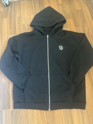 Suvene - black hoodie - Säljer en svart hoodie med dragkedja och huva. Den har en vit broderad logga på bröstet och två fickor framtill. Hoodien är enkel och stilren, perfekt för en avslappnad look. Oversize fit