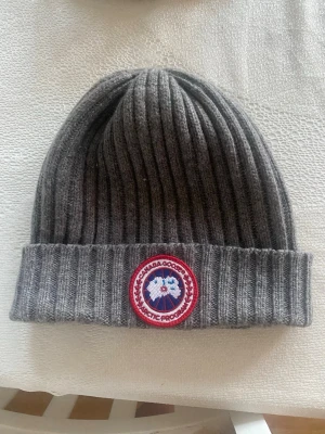Grå ribbstickad mössa Canada Goose - Snygg grå ribbstickad mössa från Canada Goose med klassisk uppvikt kant och det ikoniska runda märket framtill. Perfekt för kalla dagar och enkel att matcha med olika outfits. Mjuk och varm känsla.