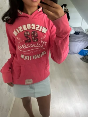 Rosa hoodie med tryck Wisconsin 32 - Säljer en rosa hoodie med vit och röd text och tryck på bröstet, där det står 'Wisconsin 32 Milwaukee College Team'. Hoodien har huva med dragsko och en stor magficka framtill. Perfekt för en avslappnad och sportig stil.