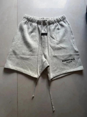 Grå Essentials Fear of God shorts - Säljer ett par ljusgrå shorts från Essentials Fear of God med snörning i midjan och tryckt logga på vänster ben. Shortsen har en relaxed passform och är tillverkade i mjuk bomull, perfekta för chill dagar.