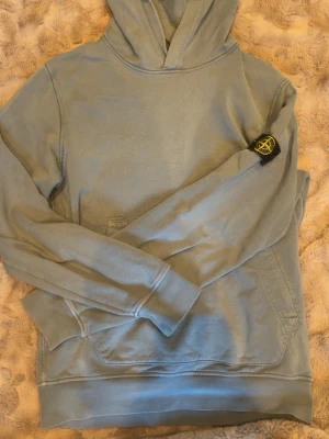 Ljusblå hoodie från Stone Island - Säljer en ljusblå hoodie från Stone Island med klassisk patch på vänster ärm. Hoodien har magficka framtill och ribbade muddar vid ärmslut och nederkant. Perfekt för dig som gillar streetwear och vill ha en stilren look.