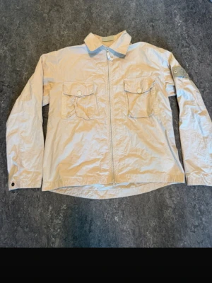 Beige overshirt från Stone Island - Snygg beige overshirt från Stone Island med dragkedja framtill och två bröstfickor med lock. Klassisk patch på vänster ärm och knappdetaljer vid ärmslut. Tillverkad i ett lätt material som passar perfekt till lager på lager.