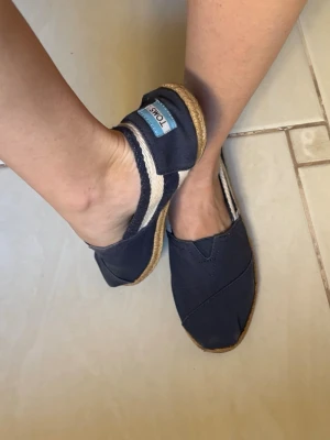 Mörkblå TOMS espadrilles - Snygga mörkblå espadrilles från TOMS med klassisk canvas-ovandel och naturfärgad sula i repimitation. Skorna har en enkel slip-on design och TOMS-logga på hälen. Perfekta för en avslappnad och trendig look.