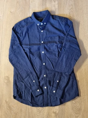 Marinblå bomullsskjorta med knappar - Snygg marinblå skjorta i bomull med klassisk krage och vita knappar framtill. Skjortan har lång ärm och en bröstficka, perfekt för en clean och stilren look. Passar dig som gillar enkel och tidlös stil. Den är väldigt skrynklig men går att platta ut med ett stryckjärn. 