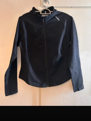 Svart träningsjacka från RACE - Snygg svart träningsjacka från RACE med hel dragkedja framtill och hög krage. Jackan har lång ärm och en minimalistisk design med logga på bröstet. Perfekt för löpning eller gymmet.
