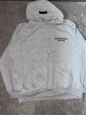 Grå hoodie Essentials Fear of God - Snygg grå hoodie från Essentials Fear of God med svart tryck på bröstet. Klassisk passform med huva och känguruficka. Perfekt för en avslappnad och trendig streetwear-look.