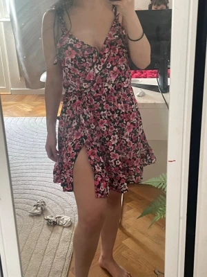 Blommig mesh kort klänning med volang - Söt kort klänning med blommigt mönster i rosa, svart och vitt. Klänningen har smala axelband, omlott framtill, volangkant och en liten slits. Perfekt för varma dagar och ger en somrig vibe.