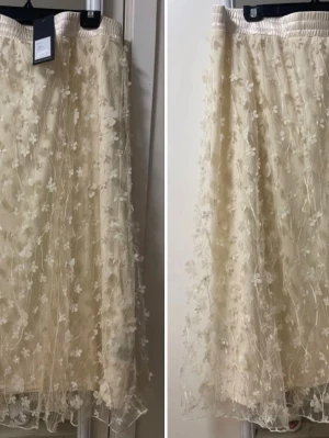 Beige långkjol med blommig spets - Romantisk beige långkjol med genomskinligt lager täckt av 3D-blommig spets och elastisk midja. Kjolen har en mjuk, flowy känsla och passar perfekt till vår och sommar för en söt och trendig look.