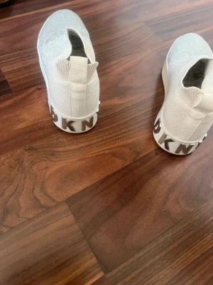 Vita slip-on sneakers från DKNY - Snygga vita slip-on sneakers från DKNY med stickad ovandel och silvriga detaljer. Skorna har en chunky sula och DKNY-logga baktill. Perfekta för dig som gillar en clean och modern stil. Materialet är textil med stretch för skön passform. Använda 2 gånger