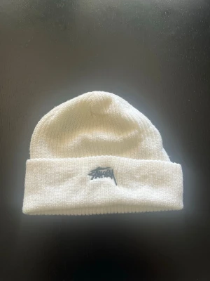 Vit ribbstickad mössa från Stussy - Säljer en stilren vit ribbstickad mössa från Stussy med uppvikt kant och broderad logga framtill. Mössan är gjord i mjukt material och passar perfekt för att hålla värmen under kalla dagar.