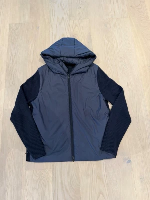 Mörkblå hybridjacka med huva - Snygg mörkblå hybridjacka med vadderad front och huva. Ärmarna är ribbstickade för extra komfort och stil. Jackan har dragkedja framtill och en minimalistisk logga på bröstet. Perfekt för kyliga dagar när du vill ha både värme och rörelsefrihet.