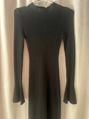 Svart ribbad långklänning - Snygg svart långklänning med ribbad struktur och långärmad design. Klänningen har en hög hals och är figurnära med lätt utsvängda ärmslut. Perfekt för dig som gillar en stilren och enkel look.