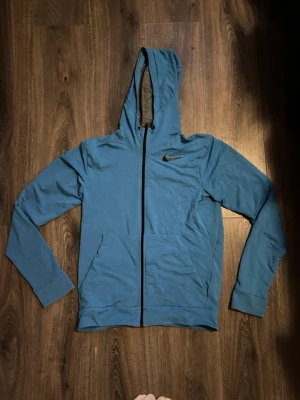 Blå Nike Dri-FIT hoodie  - Snygg blå hoodie från Nike med Dri-FIT-material som andas. Tröjan har hel dragkedja, huva och två fickor framtill. Nike-logga i svart på bröstet och mjukt, melerat foder på insidan av huvan. Perfekt för chill eller träning.