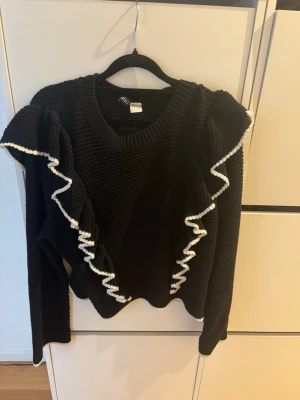 Svart stickad tröja med volang - Säljer en svart stickad tröja med långa ärmar och dekorativa volanger längs framsidan och axlarna. Volangerna har en vit kant som ger en snygg kontrast. Perfekt för dig som vill ha något extra till din outfit.