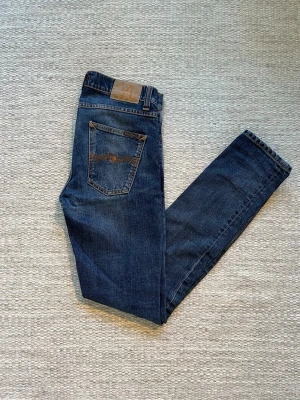 Mörkblå jeans från Nudie Jeans - Snygga mörkblå jeans från Nudie Jeans med klassisk femficksdesign och kontrastsömmar. Jeansen har raka ben och är tillverkade i slitstark denim med en skinnpatch bak i midjan. Perfekta för en avslappnad och trendig look.