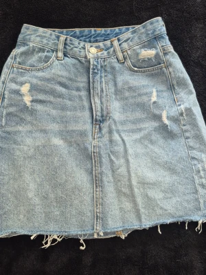 Jeanskjol från H&M - Säljer en blå jeanskjol i kort modell med slitna detaljer och rå kant nertill. Kjolen har klassiska fickor fram och bak samt bälteshällor. Perfekt för dig som gillar en avslappnad och trendig stil.Från H&M stl:34
