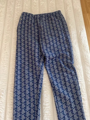 Blått loungewear-set från Lacoste - Snyggt blått loungewear-set från Lacoste med vitt geometriskt mönster. Setet består av byxor med resår i midjan och en matchande ziptröja med hög krage och dragkedja. Ikoniska Lacoste-loggan på bröstet. Perfekt för chill dagar hemma.